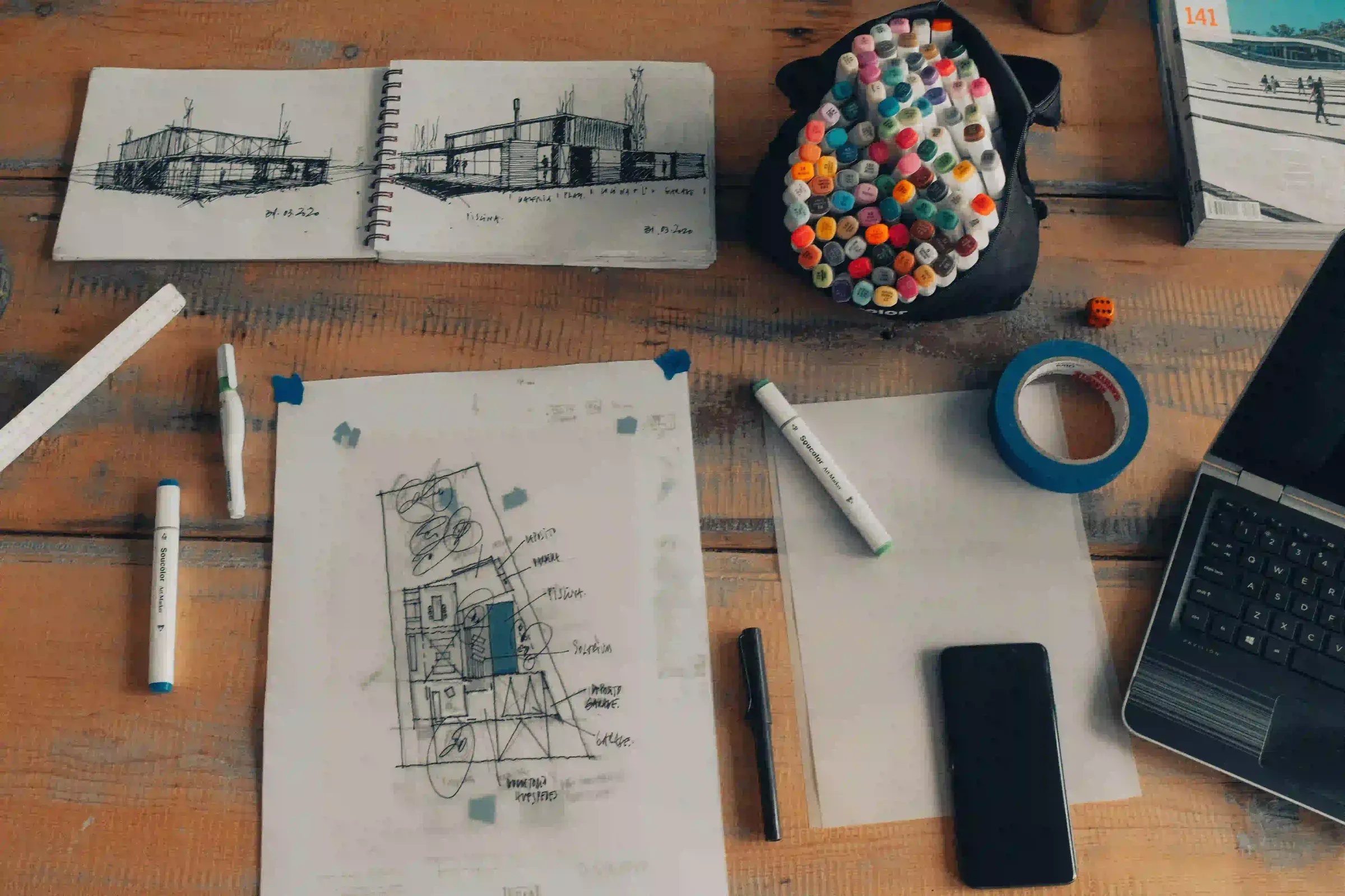 Architect's sketchbook, marker pens and a hand-drawn floor plan on a wooden desk.