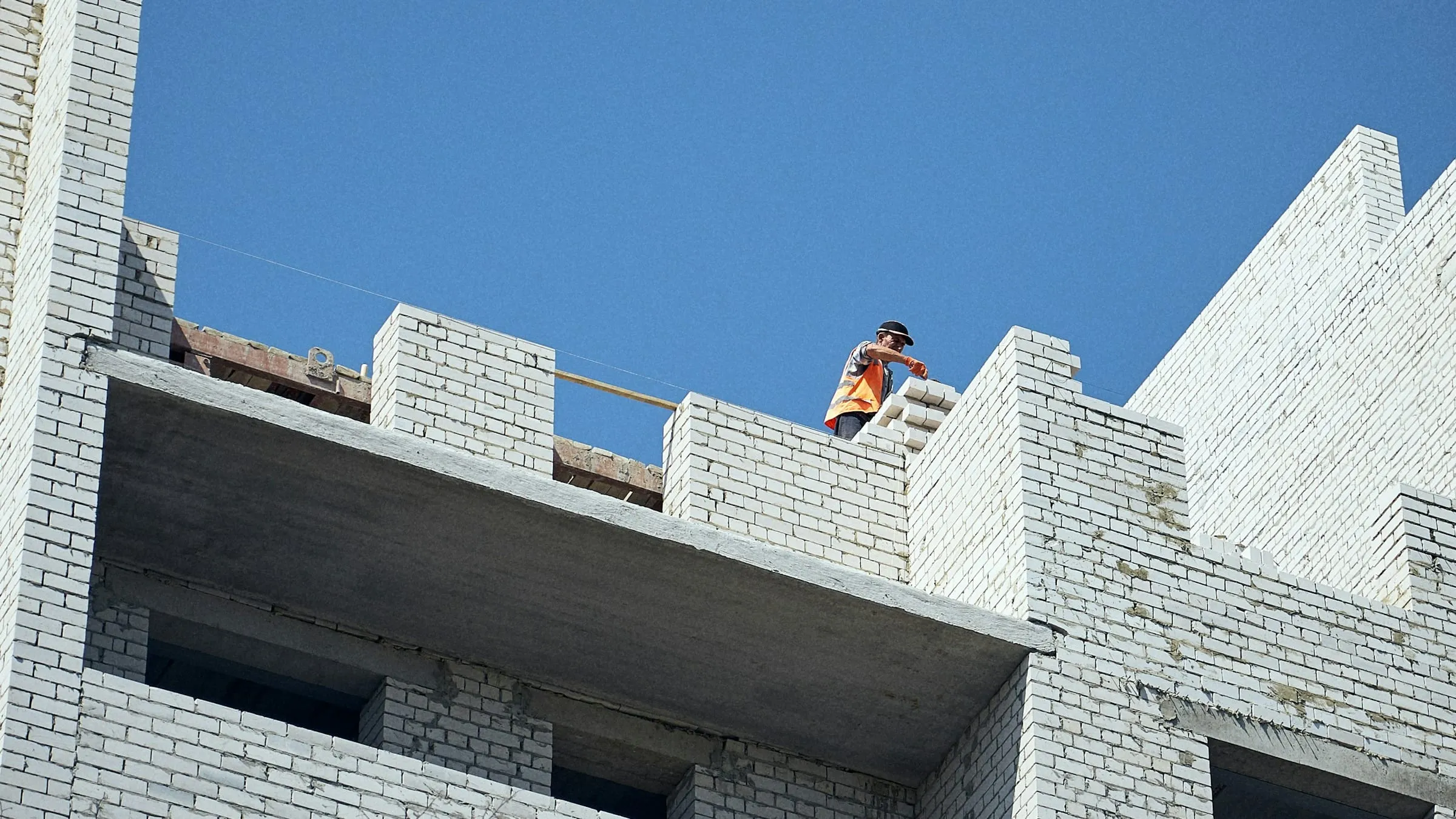 A builder at work on the roof of a new white-brick building against a clear blue sky