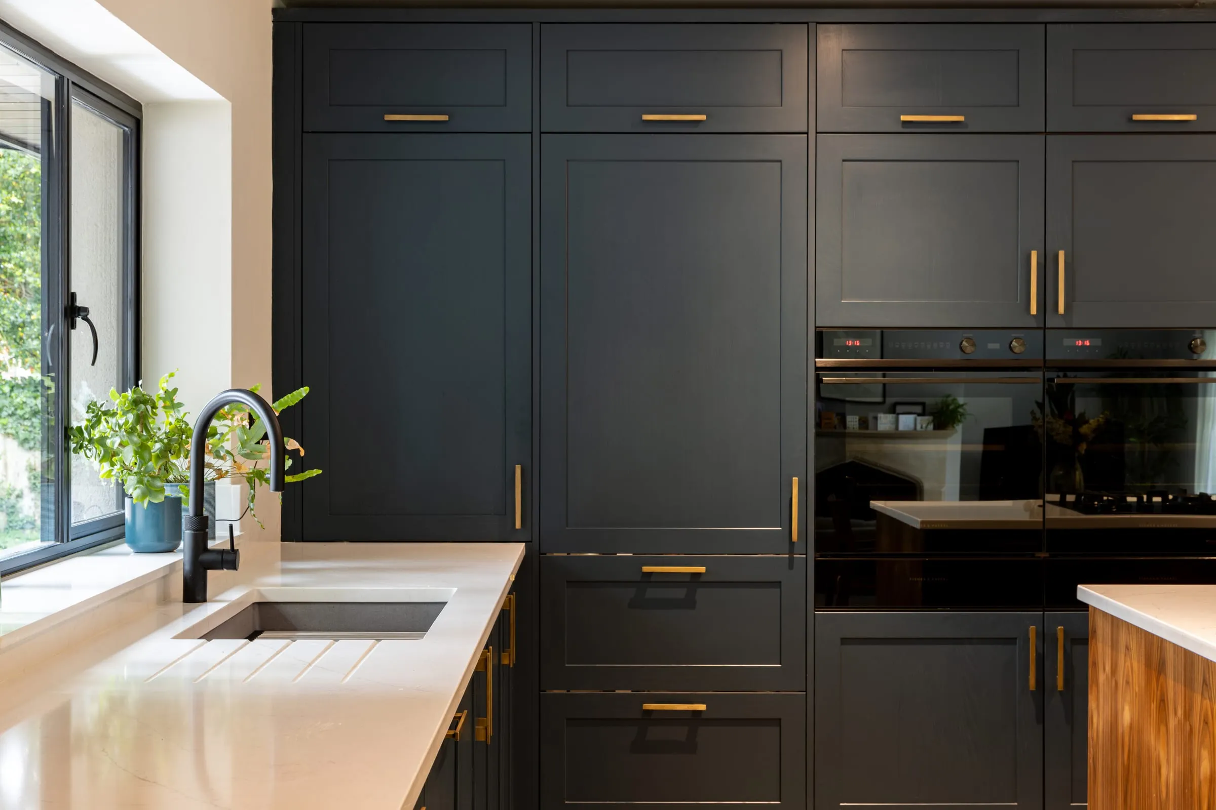 A kitchen with charcoal grey units, gold handles, double ovens and a white quartz worktop beside a window.