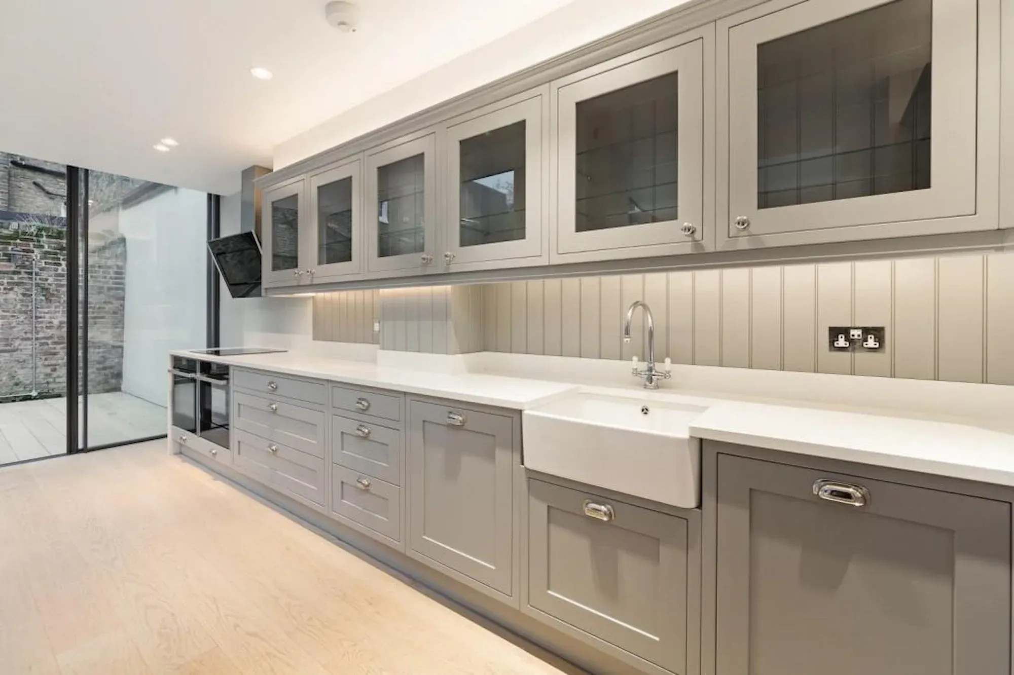 A long run of grey painted kitchen units with a butler sink, beside full-height sliding glass doors onto a brick-walled courtyard.