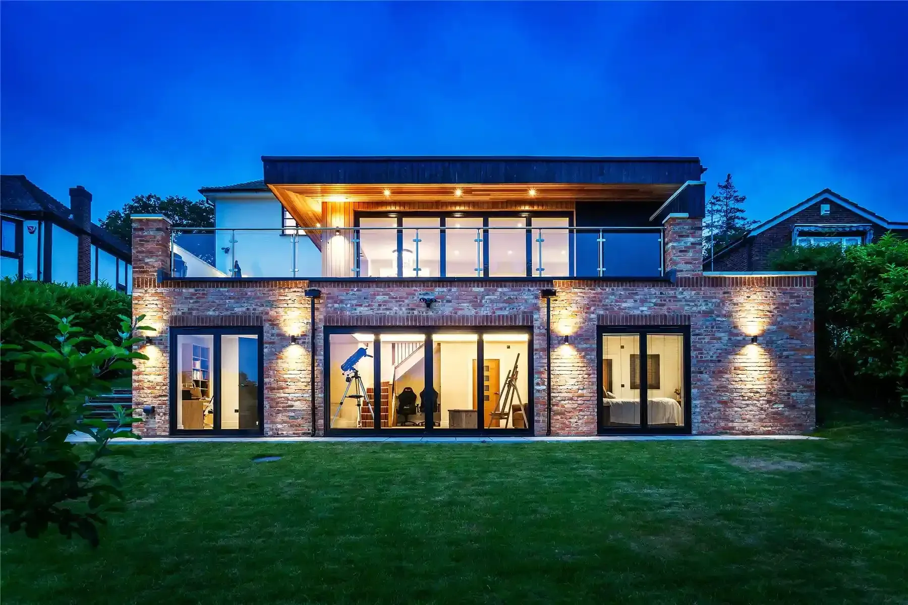 The garden side of a two-storey house at dusk, with brick below, a timber-clad upper floor and a glass balcony.