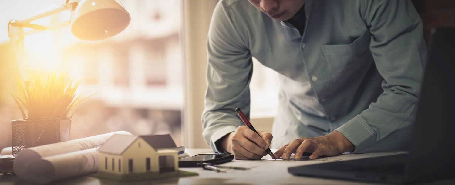 An architect leaning over a desk marking up drawings, with rolled plans and a small model house beside a desk lamp.