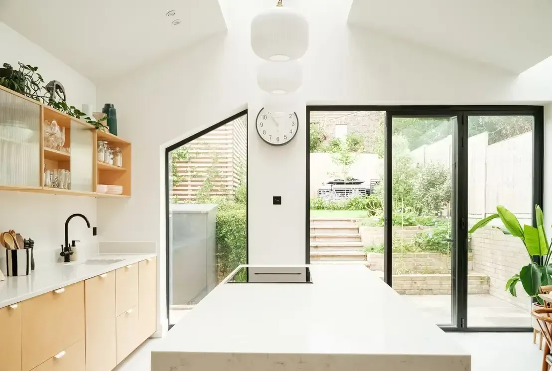 A kitchen extension with plywood units, a white island, a pitched roof with rooflights and black-framed glazed doors onto a terraced garden.