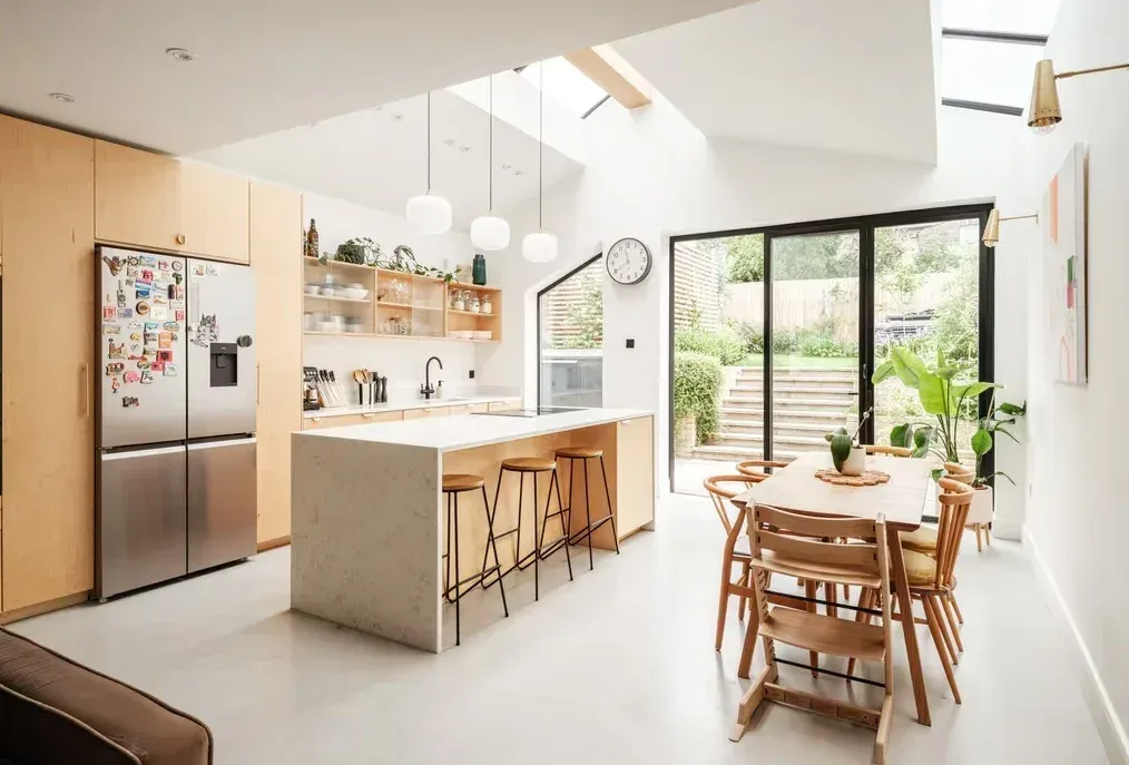 A kitchen and dining extension with plywood units, a white island, a pitched ceiling with rooflights and black-framed glazed doors onto garden steps.