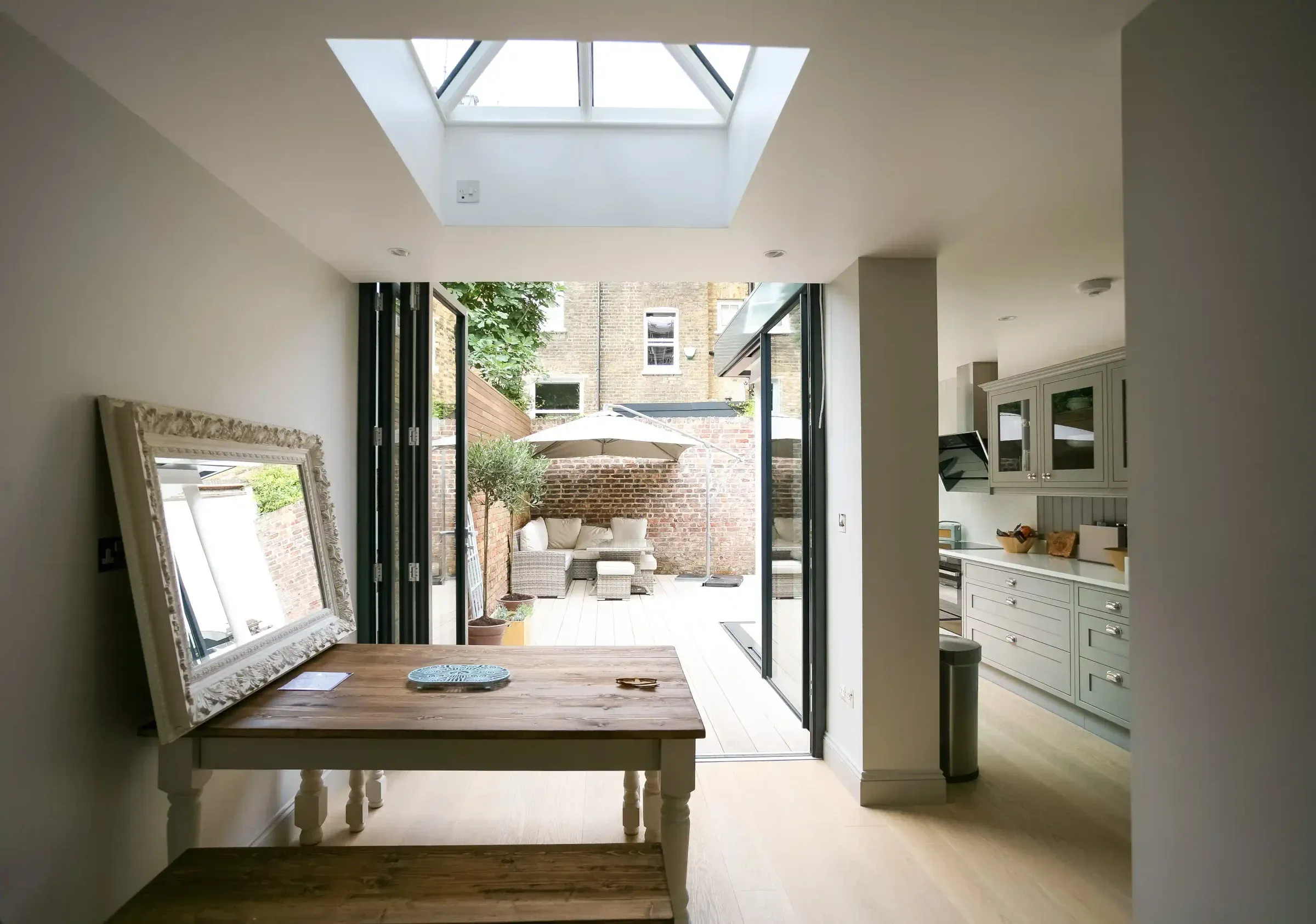A rear extension with a roof lantern over a dining table and folding doors open onto a brick-walled courtyard, with a grey kitchen to one side.