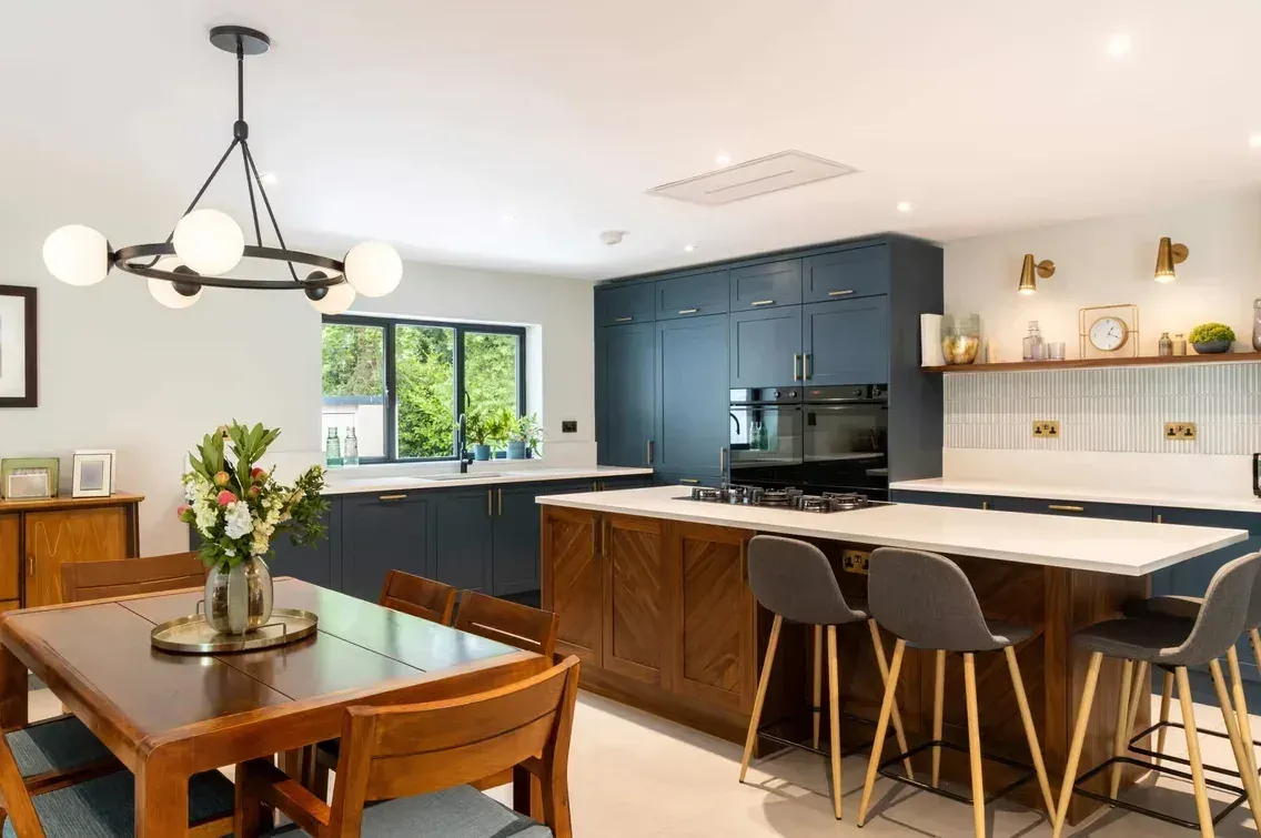 A kitchen and dining room with navy blue units, a walnut island with grey bar stools, a timber dining table and a ring-shaped globe light.