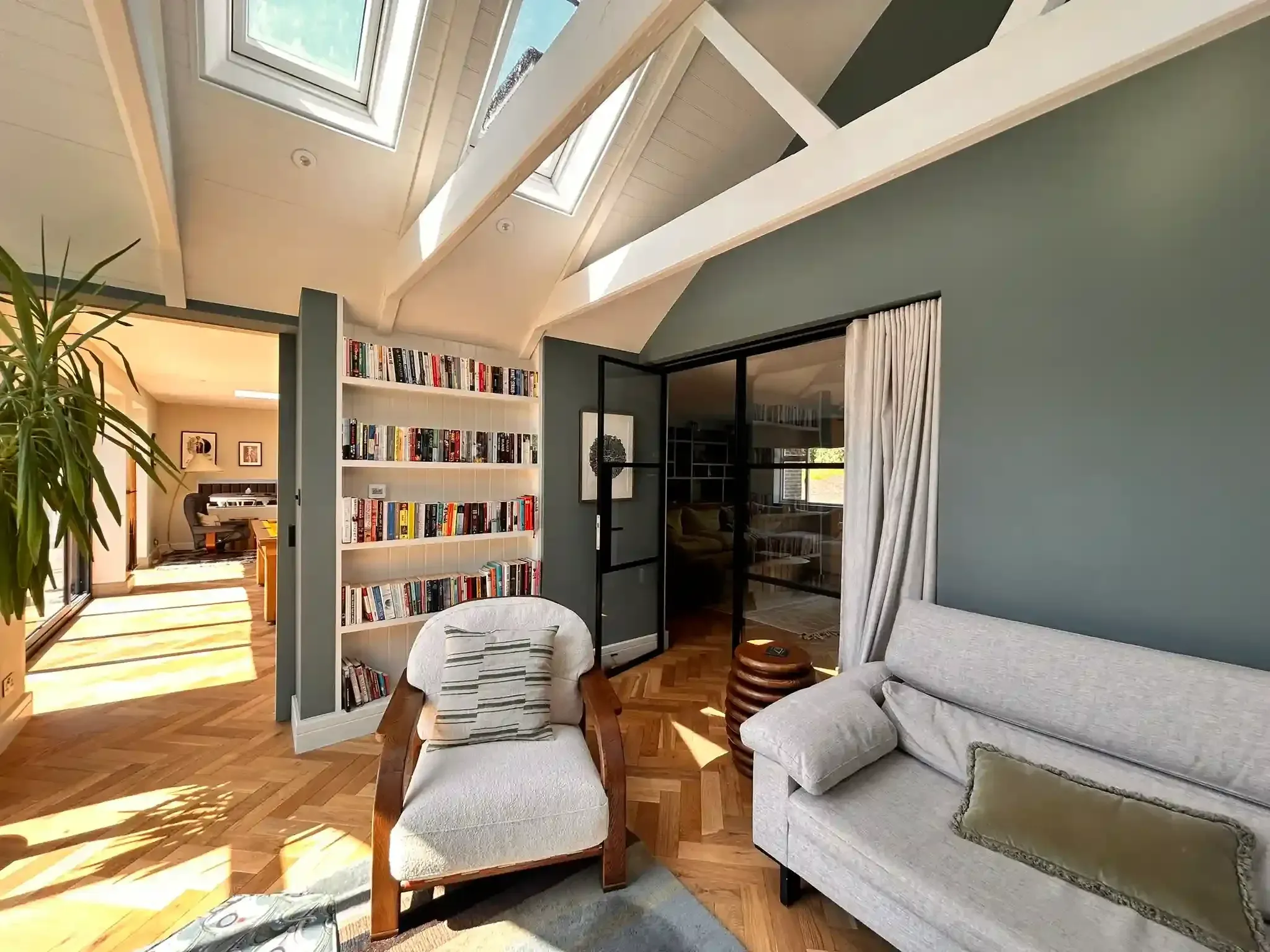 A sitting room under a vaulted ceiling with rooflights, built-in bookshelves, herringbone timber floor and glazed steel-framed doors to the next room.