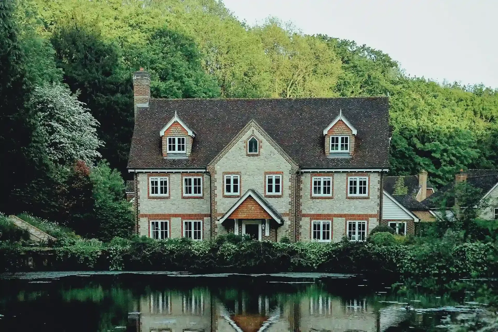 A brick and flint house with dormer windows, surrounded by trees and reflected in a pond.