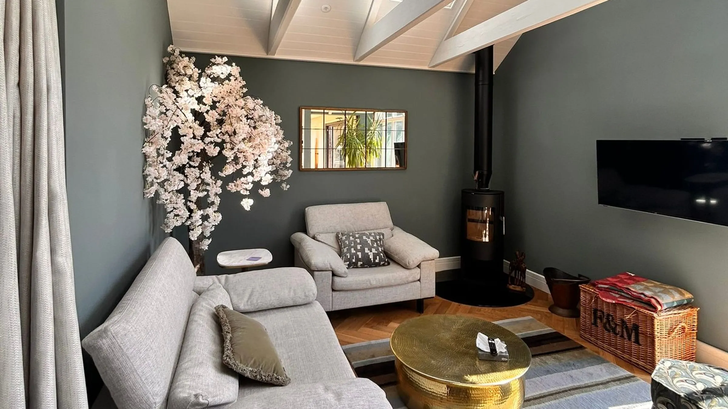 A garden room extension with grey-green walls, a sofa and armchair around a brass table, and a wood-burning stove