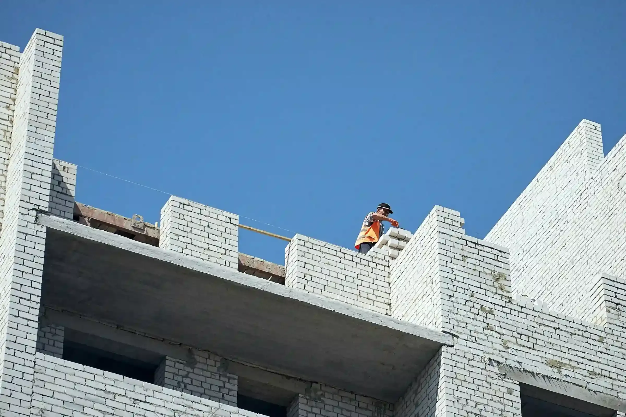 A bricklayer in a high-visibility vest working at the top of a white brick wall.