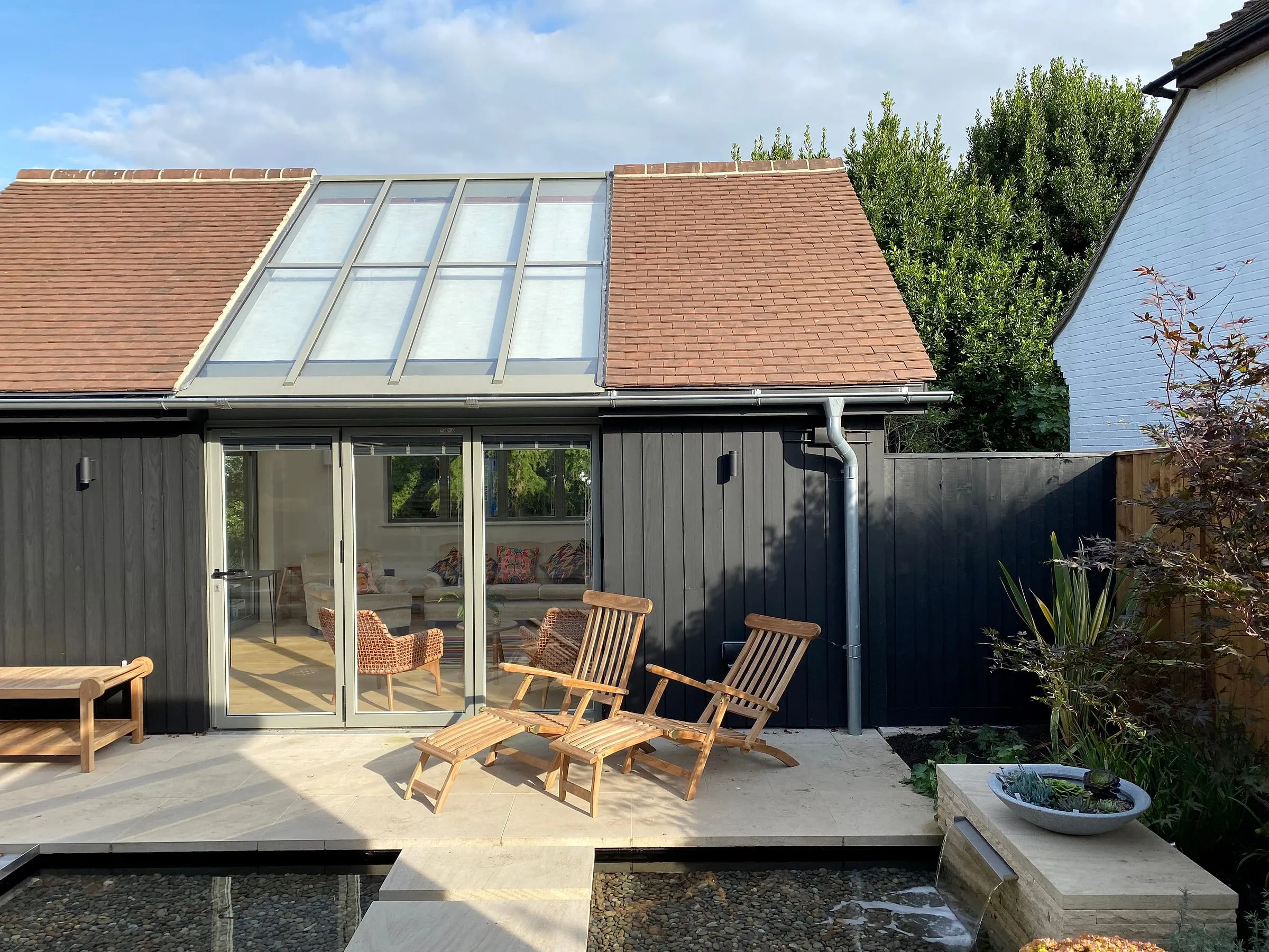 The garden room from the terrace, with a glazed roof slope, black cladding and deck chairs by the pond