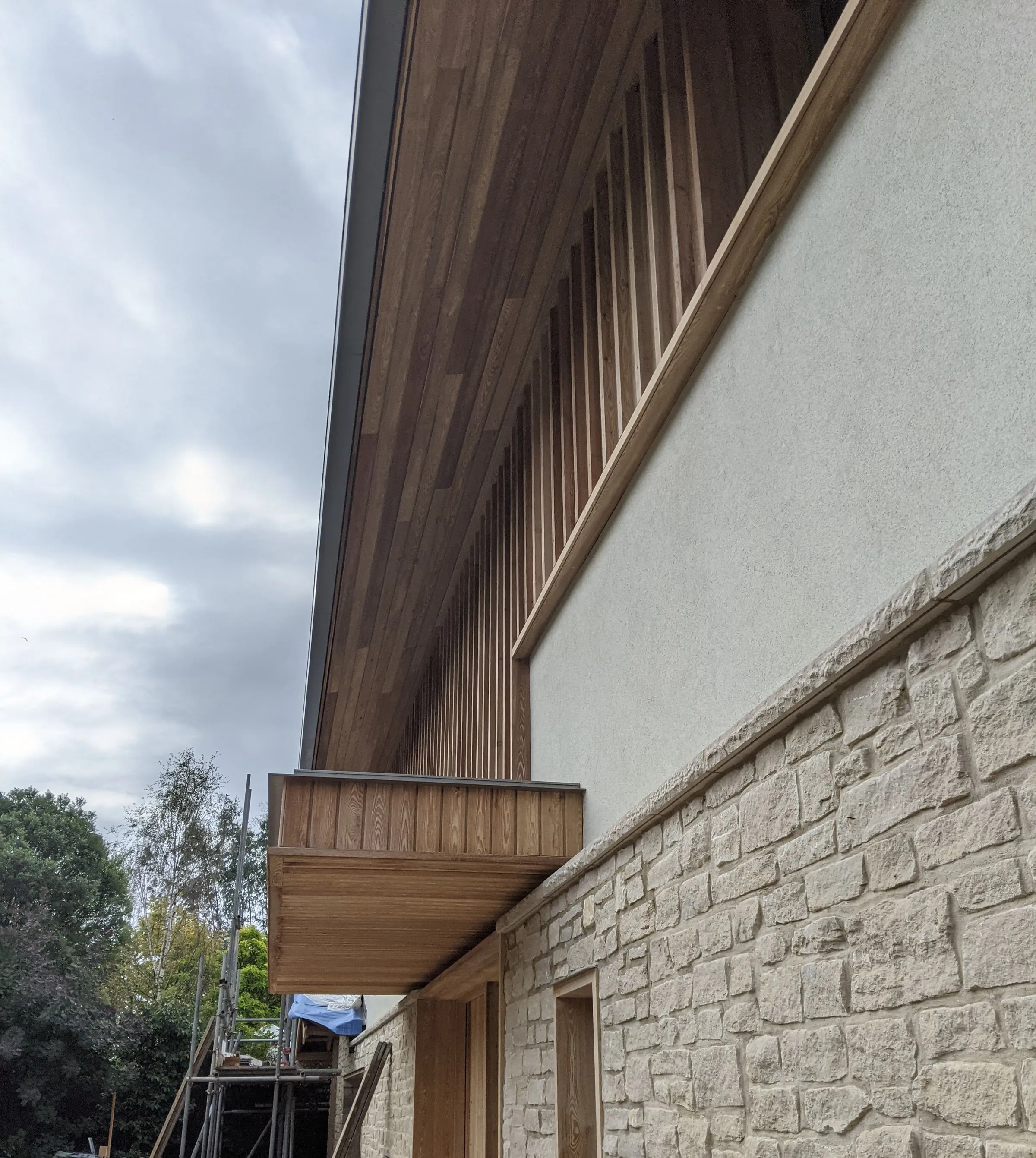 Looking along the larch soffit and fin screen above the stone plinth, with the scaffold still up beyond