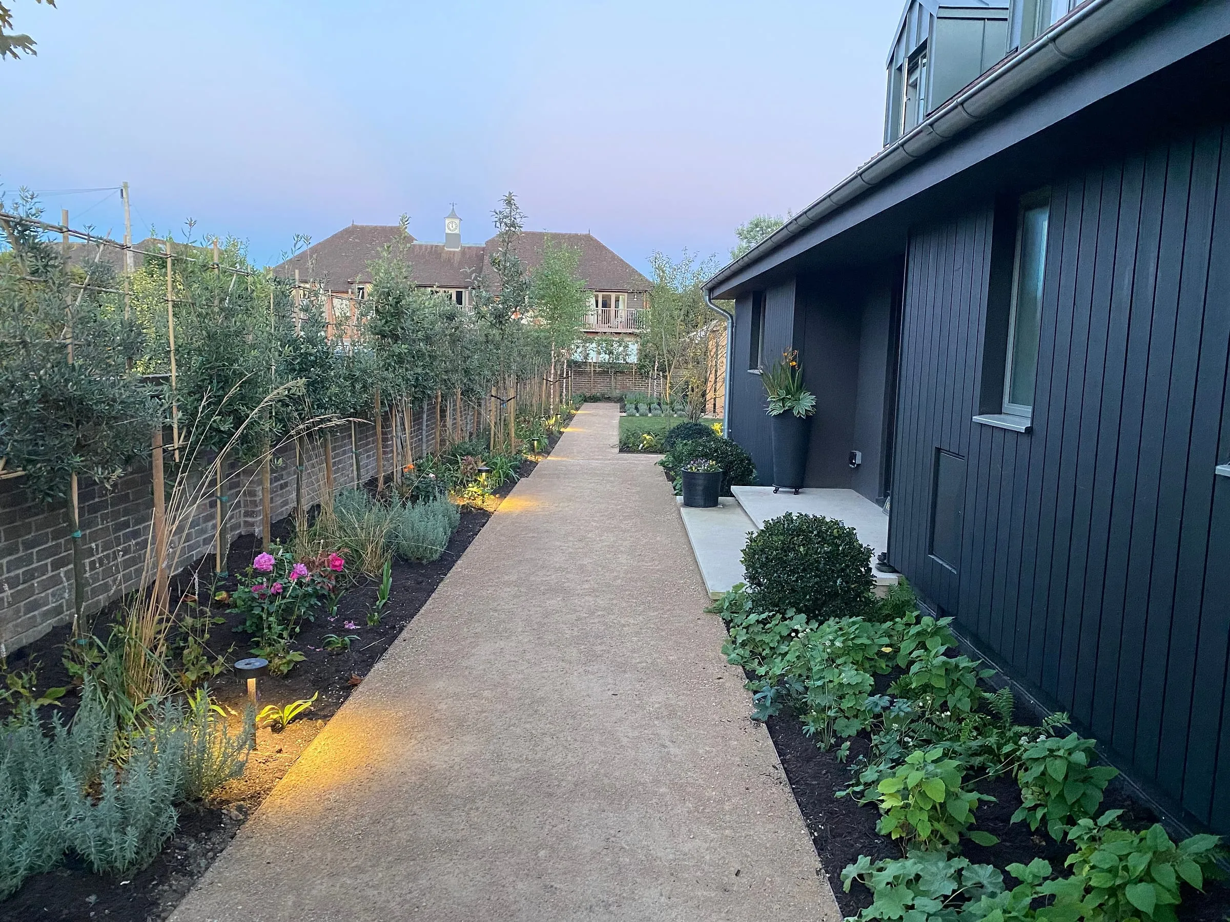 The gravel path along the side of the black-clad house at dusk, lit by low garden lights