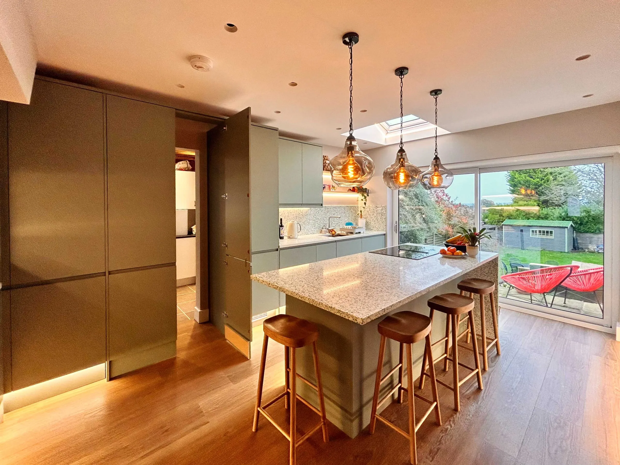 The replanned Guildford kitchen, with tall sage green units, a stone-topped island and pendant lights